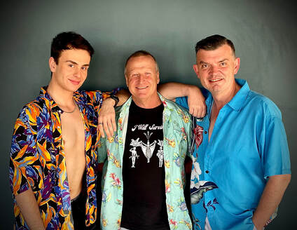 The three leads of Priscilla! The men are standing close together against a grey wall and are smiling. 